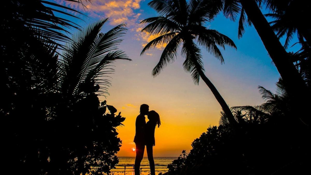 Silhouette of a couple sharing a romantic moment on a tropical beach at sunset with palm trees.