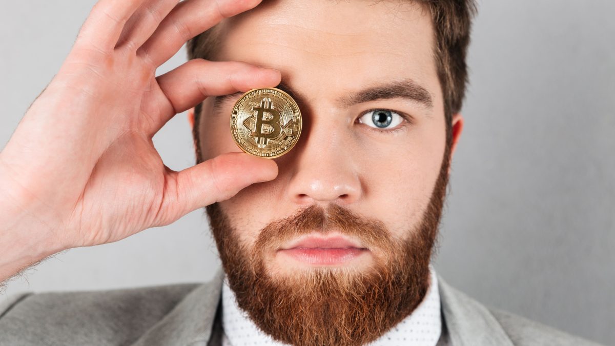 Close up of a serious businessman dressed in suit holding bitcoin at his face isolated over gray background
